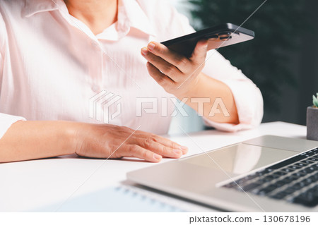 Close-up shot, woman sits at a wooden table using her smartphone, with her finger touching the phone screen as she navigates mobile banking, online shopping, and social media networks. 130678196