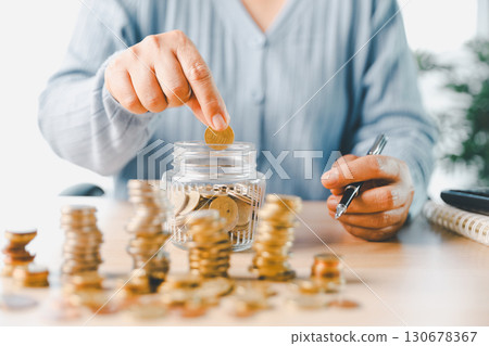 Hand places coin into saving jar, representing how smart finance, investment, banking practices turn money, cash, currency into financial wealth drives business growth and strengthens the economy. Hand places coin into saving jar, representing how smart finance, investment, banking practices turn money, cash, currency into financial wealth drives business growth and strengthens the economy. 130678367