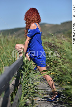 Woman with red hair in blue dress standing on wooden footbridge in a reed field 130678375
