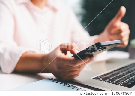 Close-up shot, woman sits at a wooden table using her smartphone, with her finger touching the phone screen as she navigates mobile banking, online shopping, and social media networks. Close-up shot, woman sits at a wooden table using her smartphone, with her finger touching the phone screen as she navigates mobile banking, online shopping, and social media networks. 130678807