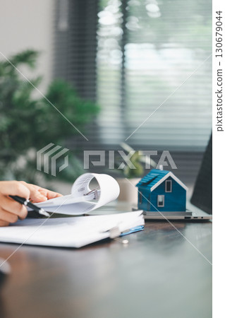 Real estate agent and customer are discussing contract to buy house, with house model in background symbolizing real estate, insurance, and loan arrangements involved in process. Real estate agent and customer are discussing contract to buy house, with house model in background symbolizing real estate, insurance, and loan arrangements involved in process. 130679044