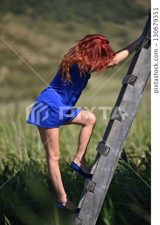 Red haired woman in vibrant blue dress and high heels ascends a rustic wooden ladder in the outdoors 130679351
