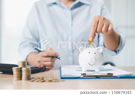 Concept of saving money and investment planning. A businesswoman drops coin into white piggy bank on office desk with documents, symbolizing financial security, budget management and future wealth. Concept of saving money and investment planning. A businesswoman drops coin into white piggy bank on office desk with documents, symbolizing financial security, budget management and future wealth. 130679381