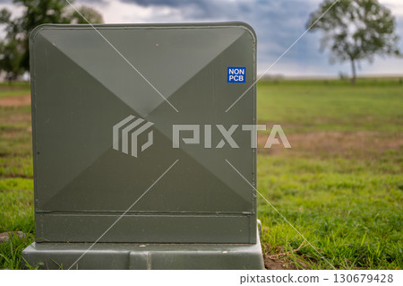 Utility box marked as non-PCB located in a green field with trees in the background during overcast weather 130679428