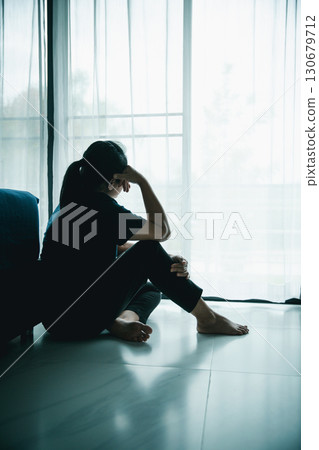 A lonely woman sits on the floor by window with hand on head in dark room. Concept of depression, sadness, anxiety, stress, mental health problem, loneliness and emotional struggle in life. 130679712