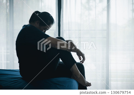 A stressed woman sitting alone on bed in dark room, holding her head down, symbolizing depression, anxiety, hopelessness, loneliness, mental health struggle, burnout and emotional pain. A stressed woman sitting alone on bed in dark room, holding her head down, symbolizing depression, anxiety, hopelessness, loneliness, mental health struggle, burnout and emotional pain. 130679754