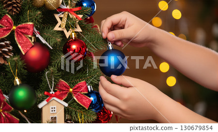 Child decorating Christmas tree with colorful baubles, holding blue ornament among red, green and gold festive decorations 130679854