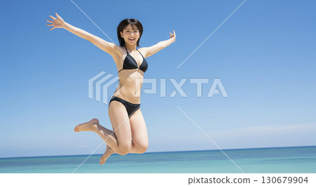 A woman in a bikini jumps energetically with her arms outstretched against the sky 130679904