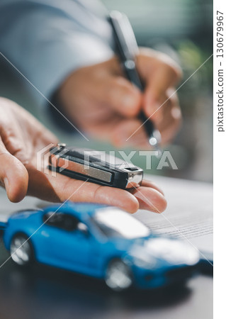 Business office, woman employee finalizes deal with client, handing over car key after signing contract and reviewing insurance and loan documents for sale or rent of vehicle at table. Business office, woman employee finalizes deal with client, handing over car key after signing contract and reviewing insurance and loan documents for sale or rent of vehicle at table. 130679967