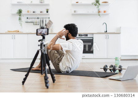 Young adult man exercising on yoga mat recording fitness video at home for blog content. Scene emphasizes health, sports, and online sharing using compact gym equipment in bright kitchen setting. Young adult man exercising on yoga mat recording fitness video at home for blog content. Scene emphasizes health, sports, and online sharing using compact gym equipment in bright kitchen setting. 130680314