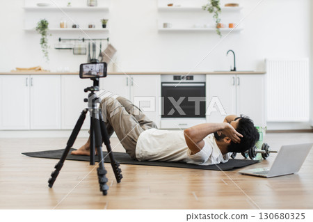 Man recording video blog on fitness and healthy living performing abs exercises, showcasing equipment, camera setup, modern kitchen environment for engaging content creation, features indoor workout 130680325