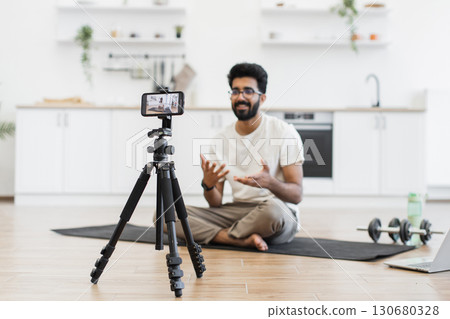 Adult man sitting on mat recording video about fitness, exercise benefits. Engaging vlog activity promoting health and workout. Explaining exercises suitable for environments during recording session. 130680328