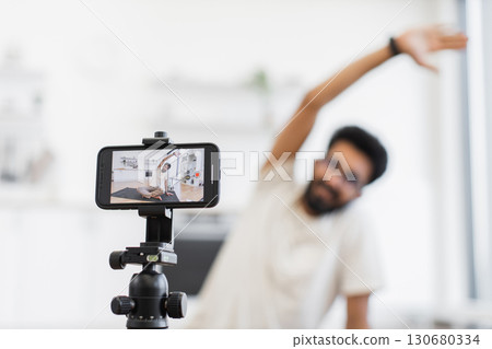 Young adult man records fitness tutorial at home using smartphone camera, focusing on arm stretches. Promotes health, fitness, and active lifestyle through shared video content on personal platforms 130680334
