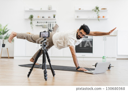 Man showcasing yoga pose during online blogging session in home kitchen, maintaining balance. Middle-aged male focused on fitness, engaging audience virtually emphasizing healthy lifestyle 130680368