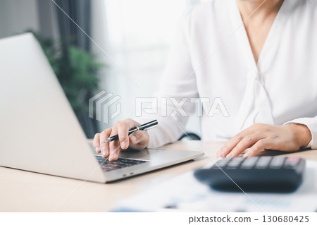 Businesswoman works on laptop while holding pen and reviewing financial documents at office desk with calculator. Concept of accounting, business planning, financial analysis and productivity. 130680425