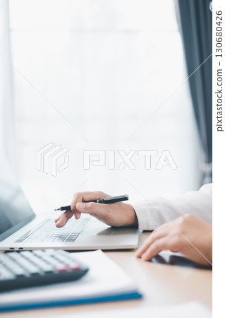 Businesswoman works on laptop while holding pen and reviewing financial documents at office desk with calculator. Concept of accounting, business planning, financial analysis and productivity. 130680426
