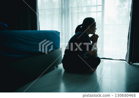 A woman sitting on the floor near window with hands clasped in thoughtful pose. Concept of depression, loneliness, anxiety, prayer, hope, mental health, emotional struggle and self-reflection. A woman sitting on the floor near window with hands clasped in thoughtful pose. Concept of depression, loneliness, anxiety, prayer, hope, mental health, emotional struggle and self-reflection. 130680468