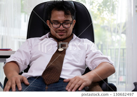 Exhausted businessman in office chair loosening tie and resting after work, symbolizing stress relief, burnout, fatigue, workplace pressure, overwork, and lifestyle challenges 130682497
