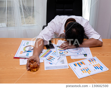 Exhausted businessman in office resting head on desk with documents and holding glass of whiskey, symbolizing burnout, financial stress, overwork, unhealthy lifestyle, alcohol addiction, and corporate 130682500