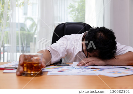 Overworked businessman in office with head on desk holding whiskey glass among financial documents, symbolizing burnout, alcohol abuse, workplace stress, financial crisis, and unhealthy lifestyle Overworked businessman in office with head on desk holding whiskey glass among financial documents, symbolizing burnout, alcohol abuse, workplace stress, financial crisis, and unhealthy lifestyle 130682501
