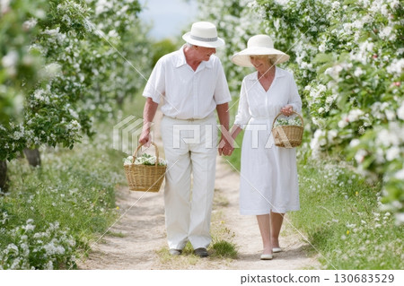 Senior couple walking through a blooming orchard holding hands and carrying baskets of white flowers, dressed in white clothes and sun hats on a sunny day Senior couple walking through a blooming orchard holding hands and carrying baskets of white flowers, dressed in white clothes and sun hats on a sunny day 130683529