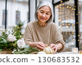 An elderly woman smiles gently while arranging small pumpkins and flowers at a cozy, festive table indoors, surrounded by warm holiday lights An elderly woman smiles gently while arranging small pumpkins and flowers at a cozy, festive table indoors, surrounded by warm holiday lights 130683532