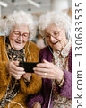 Two elderly women with white curly hair, wearing headphones and colorful sweaters, smiling and watching something on a smartphone together Two elderly women with white curly hair, wearing headphones and colorful sweaters, smiling and watching something on a smartphone together 130683535