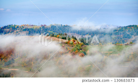 Picturesque view of hillside adorned with vibrant autumn foliage, ranging from rich greens to fiery reds and golden yellows. Wisps of fog drift across landscape under bright blue sky. 130683873
