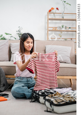 A young woman sits on the floor packing clothes into a suitcase in a bright, modern living room with plants and stylish decor. A young woman sits on the floor packing clothes into a suitcase in a bright, modern living room with plants and stylish decor. 130684833