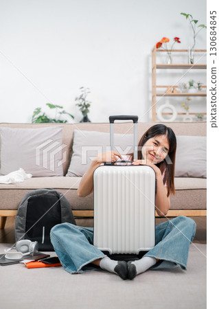 Smiling woman sitting with suitcase in cozy living room, preparing for a travel journey. Modern lifestyle and adventure concept. 130684845