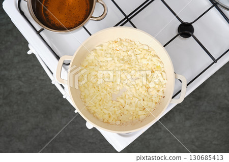 Chopped onions cooking in a pan on a stove with simmering pot of broth nearby Chopped onions cooking in a pan on a stove with simmering pot of broth nearby 130685413