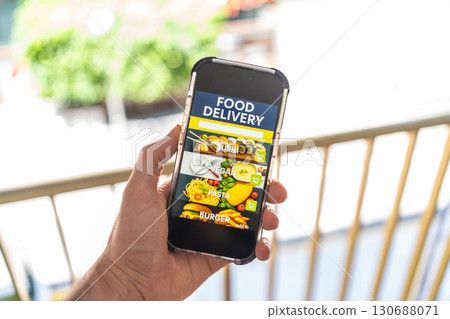 Hand holding smartphone with food delivery app interface on screen, showing sushi, vegan, pasta and burger categories. 130688071