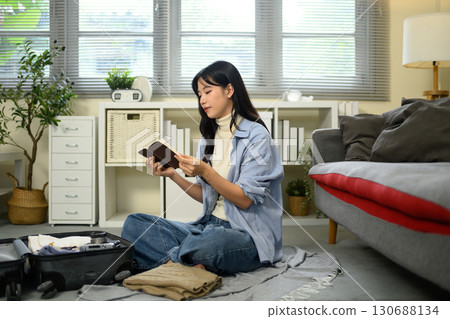 Young woman reading a travel journal while packing clothes and essentials into a suitcase Young woman reading a travel journal while packing clothes and essentials into a suitcase 130688134