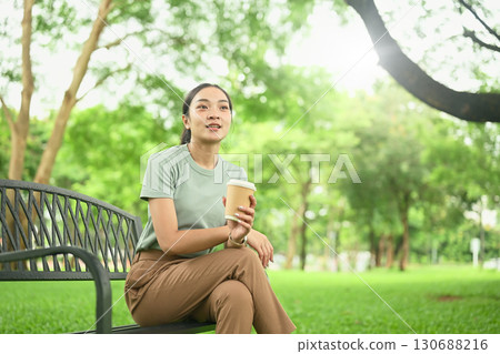 Young woman sitting on a park bench with a takeaway coffee cup, enjoying a peaceful outdoor moment 130688216