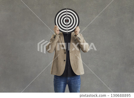 Unknown businessman hid his face behind a darts board while standing on a gray background. 130688895