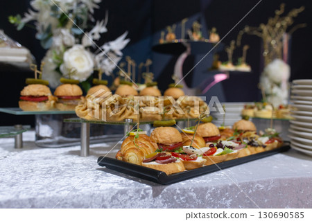Delicate mini burgers, croissants, and canapes on a banquet table. Stylish appetizer display Delicate mini burgers, croissants, and canapes on a banquet table. Stylish appetizer display 130690585