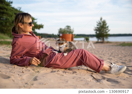 Relaxed woman lounging on sunny beach with uneasy cat curled on lap. Feline travel adaptation 130690917