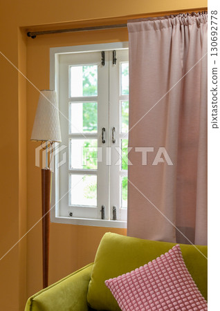 Cozy living room interior design featuring warm yellow wall and white window. pink curtain, floor lamp, green sofa, and comfortable cushion create relaxing home decor scene 130692778