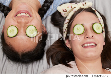 Friends, facial and skincare with cucumber by women relax, spa day and bonding while lying on a bed together. Beauty, girl and skin treatment with fruit, product and organic face mask in their home Friends, facial and skincare with cucumber by women relax, spa day and bonding while lying on a bed together. Beauty, girl and skin treatment with fruit, product and organic face mask in their home 130693667