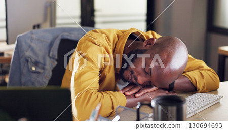 Burnout, sleeping and tired black man in the office with fatigue for working overtime on project. Exhausted, overworked and young African male designer taking a nap on his desk in the workplace. 130694693