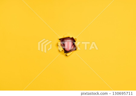 Close-up of human showing tongue through torn yellow paper 130695711