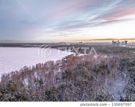 Snow-covered forest on lake shore with the first ice at sunset and the city on horizon, aerial view Snow-covered forest on lake shore with the first ice at sunset and the city on horizon, aerial view 130697997