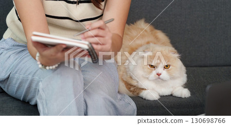 Woman writing in notebook with cat nearby, enjoying home comfort Woman writing in notebook with cat nearby, enjoying home comfort 130698766