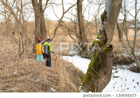 Two children explore a winter landscape by a quiet stream in the early afternoon light, surrounded by bare trees and nature's serene beauty in a peaceful countryside setting Two children explore a winter landscape by a quiet stream in the early afternoon light, surrounded by bare trees and nature's serene beauty in a peaceful countryside setting 130698855