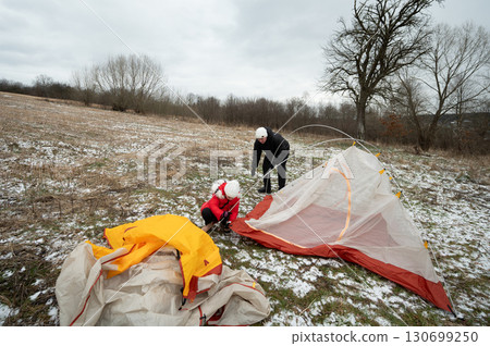 Setting up a cozy winter campsite on a chilly day in an open field surrounded by bare trees and gentle snowflakes drifting down from the gray sky 130699250