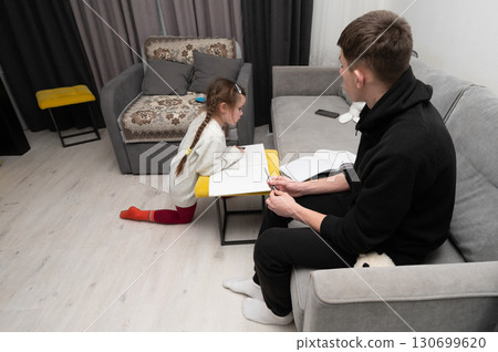 In the warm and inviting ambiance of their cozy living room during the evening hours, a child immerses themselves in the art of creative drawing, while the father watches over with attentive interest. In the warm and inviting ambiance of their cozy living room during the evening hours, a child immerses themselves in the art of creative drawing, while the father watches over with attentive interest. 130699620