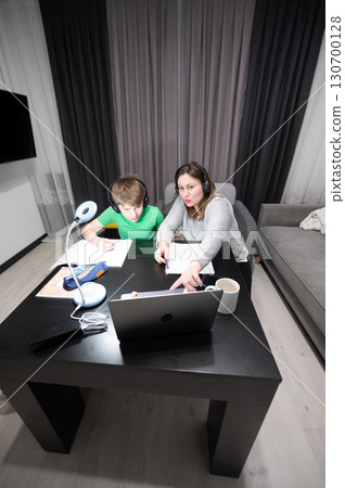 Mother and son engaged in online learning at home during evening hours, sharing knowledge and creativity in a modern living space 130700128