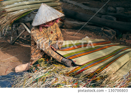 Vietnamese old female worker in traditional straw hat Non La works at reed mat factory in craft village in Vietnam Vietnamese old female worker in traditional straw hat Non La works at reed mat factory in craft village in Vietnam 130700803