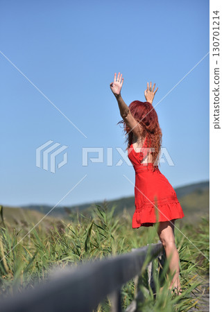 Woman with red hair in red dress walking through scenic natural wetland pathway Woman with red hair in red dress walking through scenic natural wetland pathway 130701214