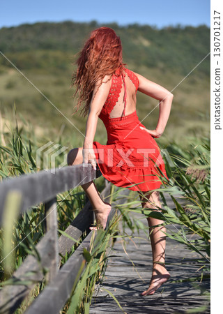 Woman with red hair in red dress walking through scenic natural wetland pathway Woman with red hair in red dress walking through scenic natural wetland pathway 130701217